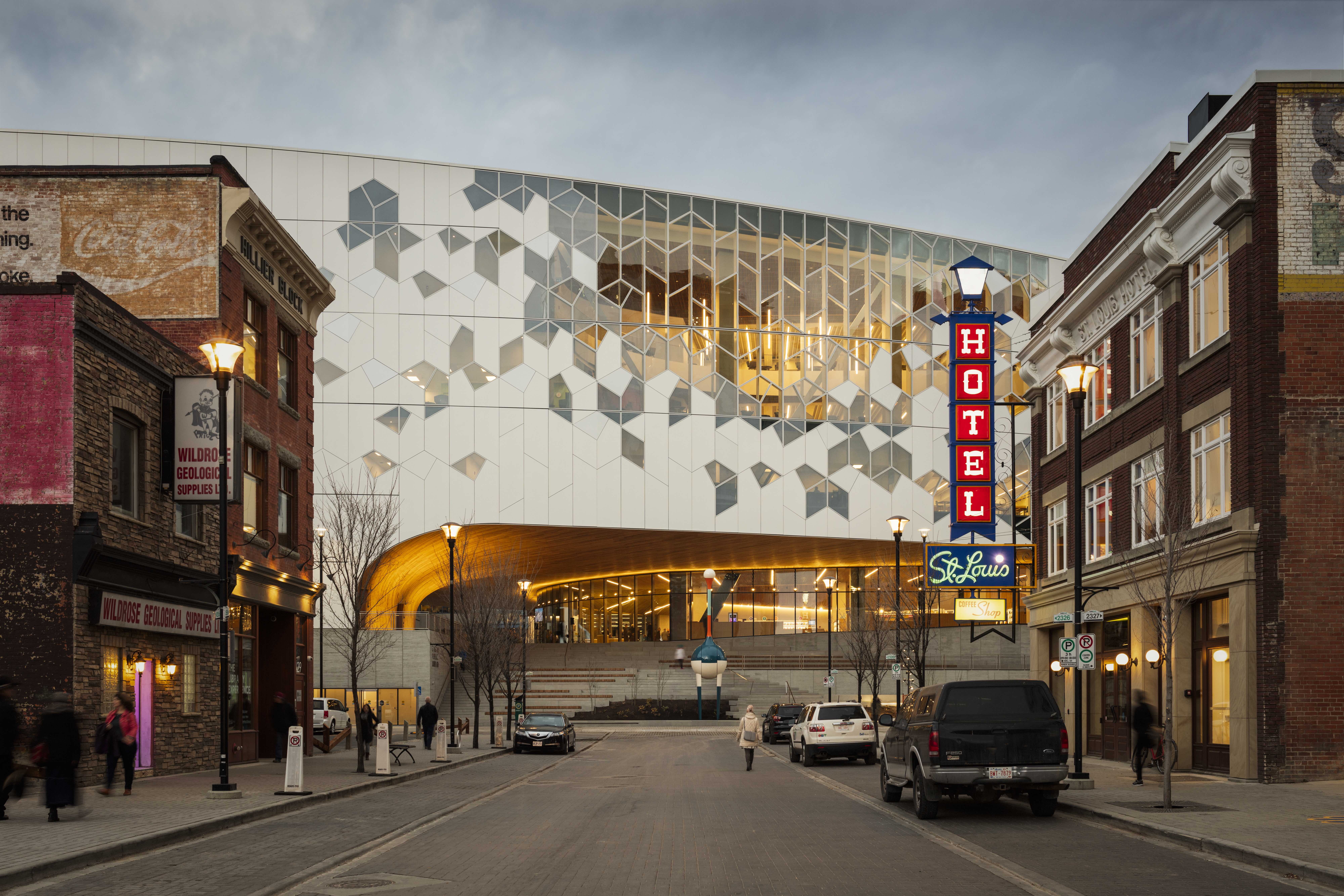 Modern architectural building with a geometric facade in the background, featuring warm lighting and a large entrance, flanked by historical buildings and street lamps in the foreground.