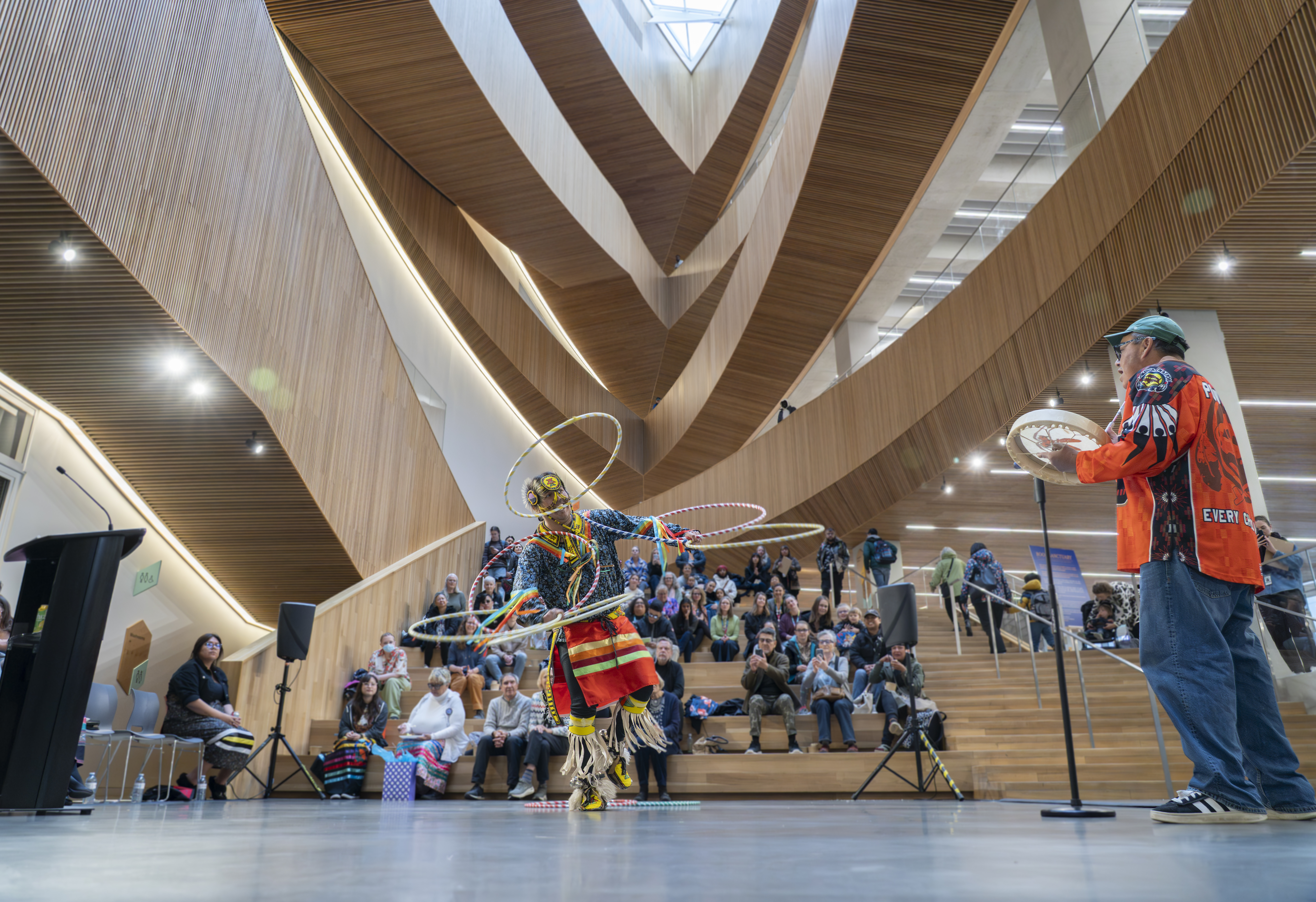 An Indigenous performer in traditional attire twirls multiple hoops during a dance performance, while a drummer plays nearby. The modern interior of a building with wooden features and an audience visible in the background.