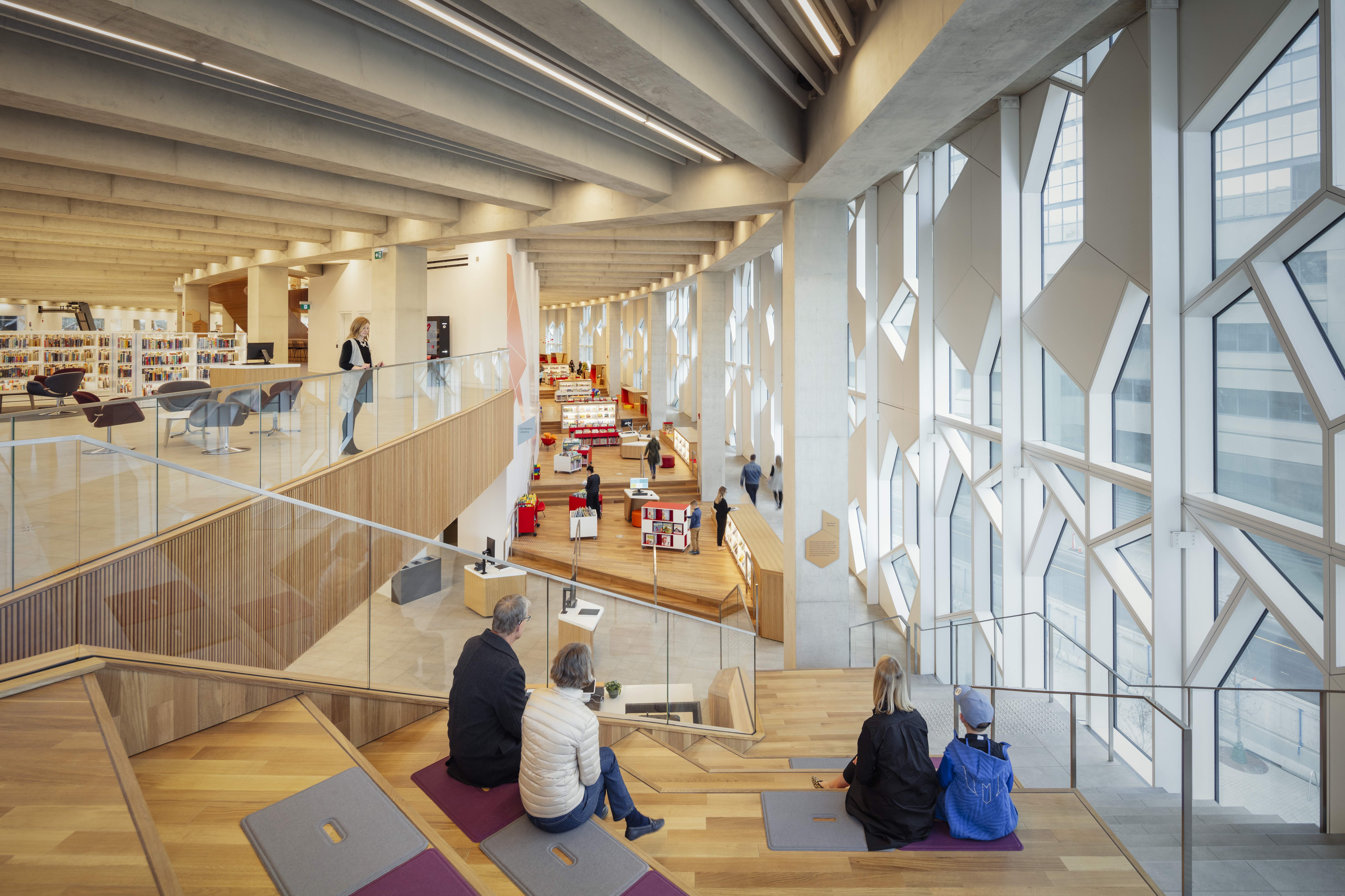 Interior view of a modern library featuring wooden stairs, large windows with geometric designs, and people engaging in various activities.