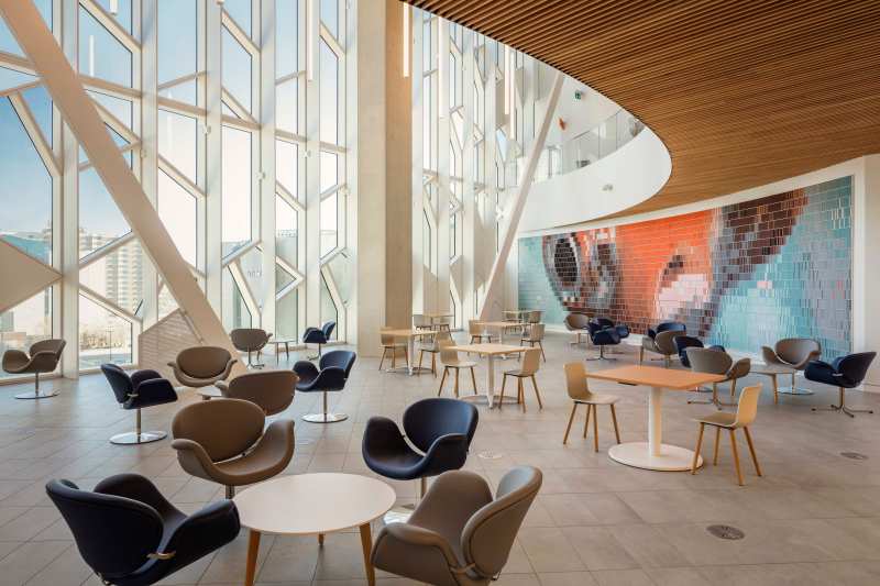 Modern interior of a bright cafe featuring a variety of seating arrangements, large windows with geometric patterns, and an artistic wall mural.