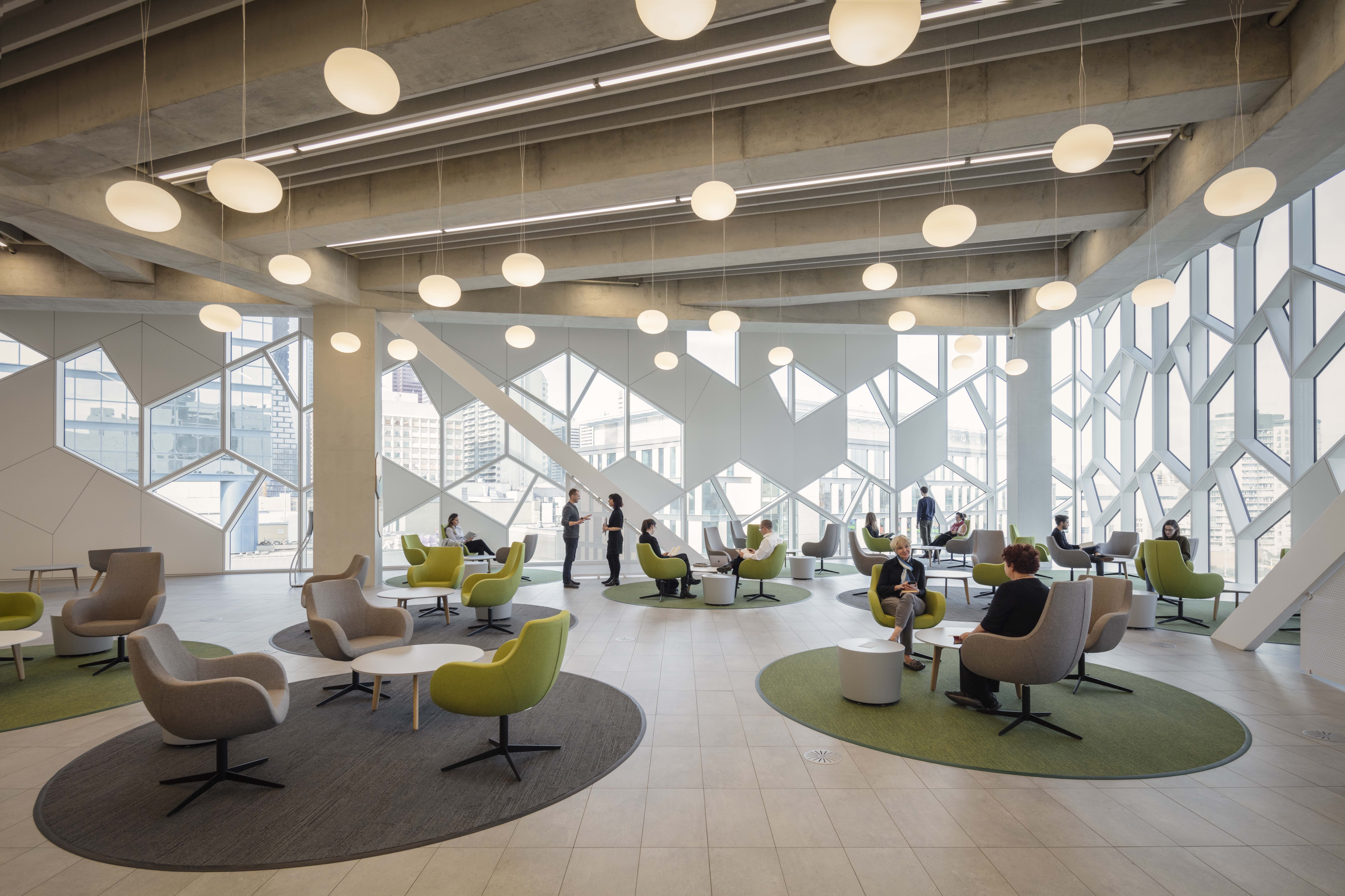 A modern, brightly lit office space featuring several comfortable lounge chairs and small tables. People are engaged in conversation, with large windows showcasing a city view and unique geometric patterns on the walls and ceiling.
