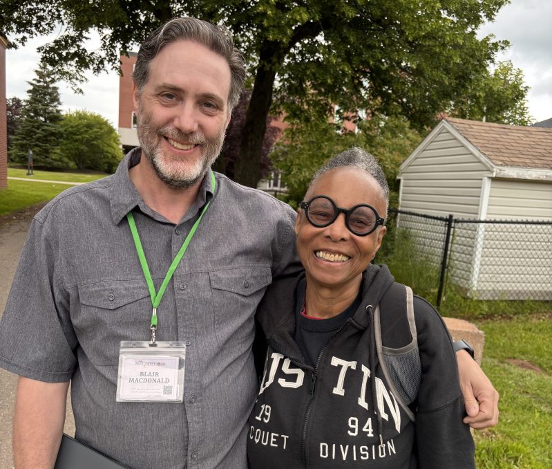 Two smiling individuals standing together outdoors, with trees and a building in the background. One person is wearing a gray shirt and has a name tag, while the other is wearing a black hoodie with 'AUSTIN' printed on it.