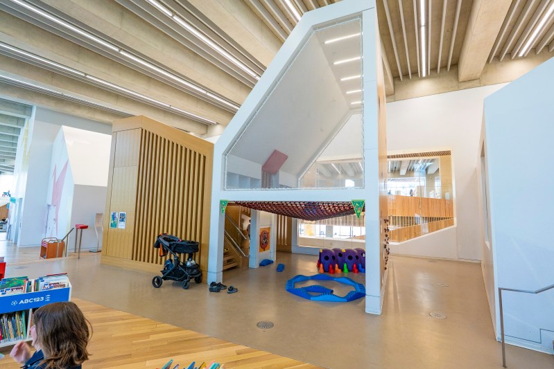 A modern library interior featuring a unique play structure with a slide and climbing elements. The area includes a children's section with colorful toys and bookshelves, designed for educational and recreational activities.