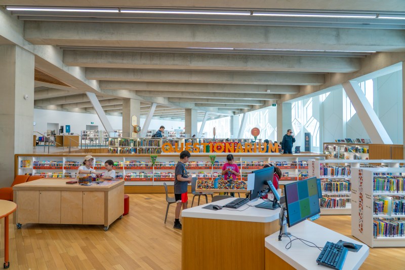 Interior view of a modern library with the word 'QUESTIONARIUM' prominently displayed. Several children are engaged in various activities, including building and exploring, while adults are browsing bookshelves in the background.