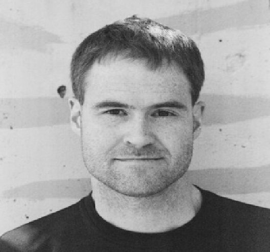 Black and white portrait of a man with short hair, wearing a black shirt, smiling softly against a textured background.