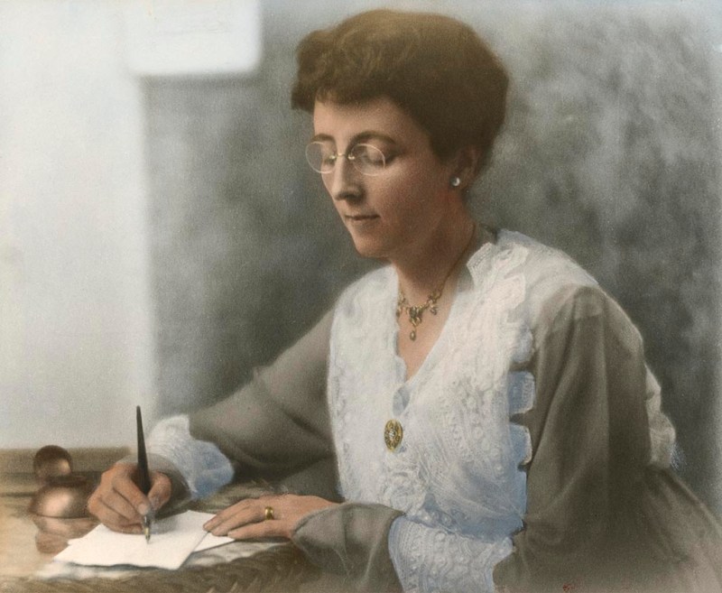 A woman with glasses is sitting at a table, writing on a piece of paper with a pen. She is wearing a light-colored blouse with lace detailing and is adorned with jewelry. The background is softly blurred.